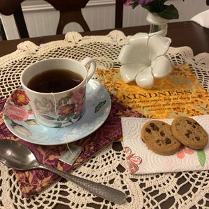 Può includere: Una tazza da tè bianca con motivi floreali piena di tè, un piattino con un motivo floreale, una bustina di tè, un cucchiaio, un libro con la parola "KINDNESS" sulla copertina, due biscotti al cioccolato con gocce di cioccolato su un tovagliolo e un fiore bianco su un tavolo con un tovagliolo all'uncinetto.