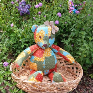 May include: A handmade patchwork teddy bear with a brown bow, sitting in a woven basket. The bear features a colorful patchwork design with red, green, blue, and yellow floral patterns. The background includes green foliage and purple flowers.