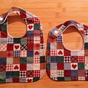 May include: Two quilted baby bibs with a patchwork design featuring red, blue, green, and cream squares with small red hearts. The bibs have a white snap closure.