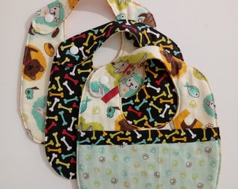 May include: Two colorful fabric baby bibs with a dog bone pattern. The bibs have a pocket on the bottom and are made of a soft, absorbent material.