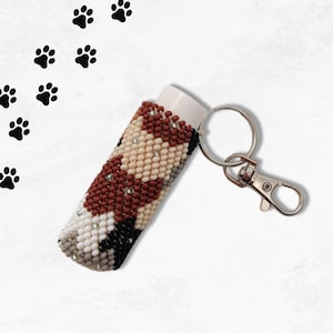 Könnte beinhalten: Ein perlenbesetzter Lippenbalsamhalter mit einem geometrischen Muster in Braun-, Weiß- und Schwarztönen. Der Halter ist an einem silbernen Schlüsselanhänger mit einem Karabinerhaken befestigt.