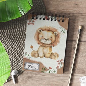 Könnte beinhalten: Ein braunes und weißes Spiralnotizbuch mit einer Aquarell-Illustration eines Löwenjungen. Das Notizbuch hat einen braunen Papierumschlag mit einem abgerissenen Rand und ein weißes Etikett mit dem Namen "Klaus" darauf.