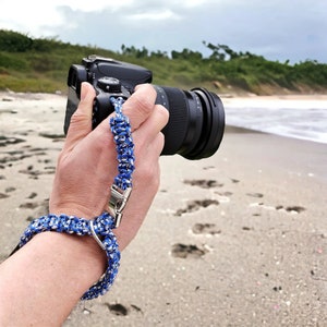 Könnte beinhalten: Ein blaues und weißes geflochtenes Kameraband mit einem silbernen Verschluss. Das Band ist an einer schwarzen Kamera mit einem Objektiv befestigt.