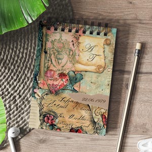 Könnte beinhalten: Ein spiralgebundenes Notizbuch mit Vintage-Design mit Blumen- und Herzmotiven. Das Cover hat einen zerrissenen Papiereffekt mit den Initialen "A T" auf einer Schriftrolle. Das Datum "22.06.1979" ist ebenfalls sichtbar. Ein Bleistift und ein Kopfhörer liegen daneben.
