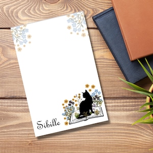 Könnte beinhalten: Ein weißer Notizblock mit einer schwarzen Katzensilhouette, die auf einem geöffneten Buch mit Blumen sitzt. Der Notizblock trägt den Schriftzug "Sibille".