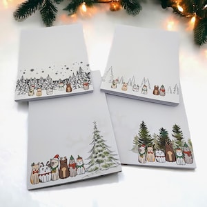 Könnte beinhalten: Vier weiße Notizblöcke mit festlichen Illustrationen. Jeder Notizblock zeigt ein anderes Design von Katzen und Bäumen, einige mit Schnee und Weihnachtsaccessoires. Die Illustrationen sind in einem skurrilen, cartoonartigen Stil gehalten.