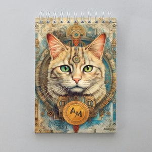 Könnte beinhalten: Ein Notizbuchcover im Vintage-Stil mit einer detaillierten Illustration einer Katze mit grünen Augen. Die Katze ist umgeben von komplizierten Mustern und Symbolen, darunter ein kreisförmiges Design mit den Buchstaben "AM" in der Mitte.