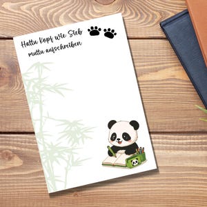 Könnte beinhalten: Ein weißer, rechteckiger Notizblock mit einer hellgrünen Bambusillustration auf der linken Seite, zwei schwarzen Pfotenabdrücken oben und einem Panda-Cartoon, der unten rechts in ein Buch schreibt. Der Text lautet: Hattu Kopf wie Sieb muttu aufschreiben.