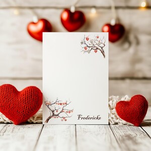 Könnte beinhalten: Eine weiße Karte mit einer handgezeichneten Illustration eines Baumes mit Herzen, die an den Ästen hängen. Die Karte trägt den Namen "Fredericke" unten. Die Karte ist umgeben von roten Strickherzen und roten Filzherzen.