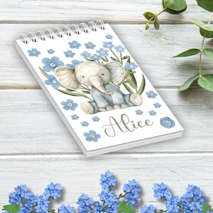 Könnte beinhalten: Ein weißes Spiralnotizbuch mit einer blauen und grünen Aquarell-Illustration eines Elefanten, der in einem Feld mit Vergissmeinnicht-Blumen sitzt. Der Name "Alice" ist in grüner Kursivschrift auf der Vorderseite des Notizbuchs geschrieben.