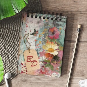 Könnte beinhalten: Spiralgebundenes Notizbuch mit Blumen- und Schmetterlingsdesign. Das Cover zeigt eine Collage aus rosa, gelben und orangen Blumen, mit einem Schmetterling und den Buchstaben "BD" auf einem Anhänger. Ein Bleistift und Ohrhörer sind ebenfalls sichtbar.