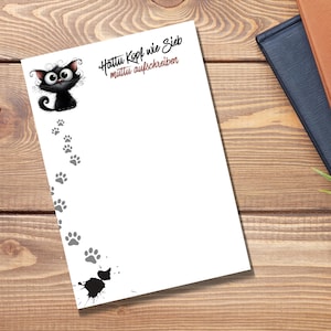Könnte beinhalten: Ein weißes Notizblatt mit einer schwarzen Cartoon-Katze mit großen Augen in der oberen linken Ecke, grauen Pfotenabdrücken, einem schwarzen Tintenklecks am unteren Rand und dem Text Hattu Kopf wie Sieb muttu aufschreiben.
