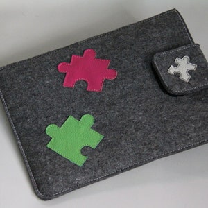 Könnte beinhalten: Eine graue Filz-Laptoptasche mit drei bunten Puzzleteilen, die auf der Vorderseite aufgenäht sind. Die Puzzleteile sind rosa, grün und weiß.