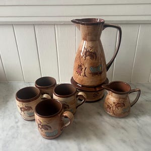 May include: A vintage ceramic pitcher and mug set with a brown and beige color scheme. The set includes a large pitcher, a creamer, and six mugs. Each piece features a decorative illustration of a covered wagon and a rider.