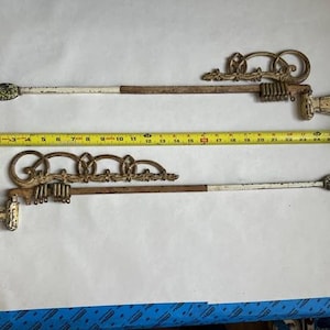 May include: Two metal curtain rods with decorative scrollwork finials, featuring a distressed white and brown finish. A yellow measuring tape is positioned between the rods, showing a length of approximately 61 cm for the metal components.