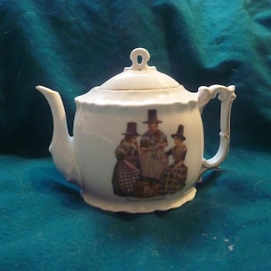 May include: White porcelain teapot with a floral design and three women in traditional Welsh dress. The teapot has a gold handle and spout.
