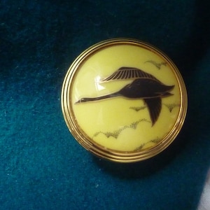 May include: A round button with a gold-toned finish, featuring a yellow background and a black silhouette of a bird in flight. The bird is flying over a stylised cloud pattern.