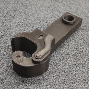Puede incluir: Una garra de plástico negro impresa en 3D con un brazo articulado y un agujero redondo en la parte superior. La garra está diseñada para ser utilizada como una herramienta o un juguete.