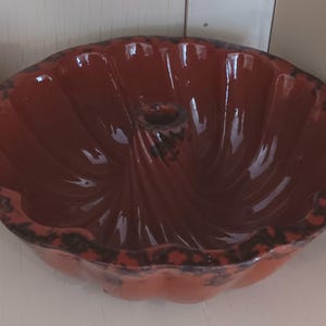 May include: A red ceramic bundt pan with a glossy finish. The pan has a fluted design and a central tube. The rim features a speckled, dark brown pattern. This baking dish is ideal for making cakes and other baked goods.