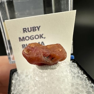 May include: A red ruby gemstone from Mogok, Burma, displayed in a small clear plastic box with white foam padding.