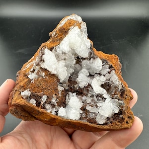 Op de afbeelding: Een ruw, natuurlijk mineraal specimen met een roestbruine matrix en clusters van witte, kristallijne formaties. Het mineraal heeft een ruw, getextureerd oppervlak. Het specimen wordt in een hand tegen een donkere achtergrond gehouden.