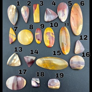 May include: Nineteen polished cabochon gemstones in various shapes including ovals, triangles, and rectangles. The stones feature swirling patterns of yellow, red, brown, and purple hues, each labeled with a white number from 1 to 19.