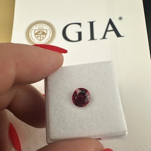 May include: A round, faceted, deep red gemstone, held in a white box. The GIA logo and text are visible in the background. The gemstone is likely a ruby or garnet, showcasing a rich, saturated color.
