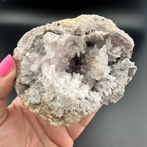 Op de afbeelding: Een geode met een ruwe, bruine buitenkant en een holte gevuld met clusters van witte en heldere kristallen. De geode wordt vastgehouden in een hand met roze nagellak tegen een zwarte achtergrond.