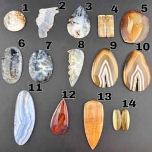 May include: A collection of fourteen polished gemstones labeled one through fourteen, featuring various shapes and patterns including banded agate, dendritic stone, and tiger eye, displayed as individual cabochons with distinct natural color variations.