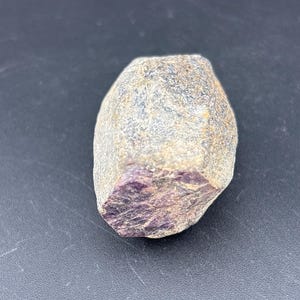 May include: A rough, natural gemstone with a mix of gray, tan, and purple hues. The stone has a textured surface and a crystalline structure visible on one side. The overall shape is irregular and rounded.