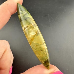 May include: A polished, elongated gemstone pendant with a pointed tip. The stone features a blend of olive green, tan, and cream colors with natural veining. The pendant is held between fingers against a dark background.