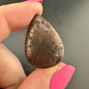 May include: A teardrop-shaped, copper-colored stone with dark spots. The stone is held between fingers with bright pink nail polish. The stone has a textured surface and a polished finish.