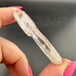Puede incluir: Un cristal claro y alargado con una textura ligeramente rugosa. El cristal se sostiene entre los dedos con esmalte de uñas rosa brillante, sobre un fondo oscuro. La forma del cristal es natural e irregular.