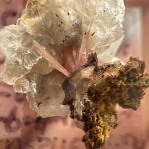 May include: A close-up of a mineral specimen featuring a cluster of clear, crystalline formations. Pink, needle-like crystals are visible at the center, surrounded by a beige and brown matrix. The specimen has a rough, textured appearance.