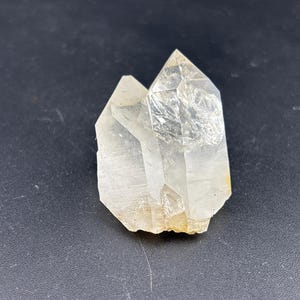 May include: A clear quartz crystal cluster with two prominent points. The crystal is translucent with visible internal structures and a slightly rough, natural texture. The base has a light, yellowish hue, set against a dark background.