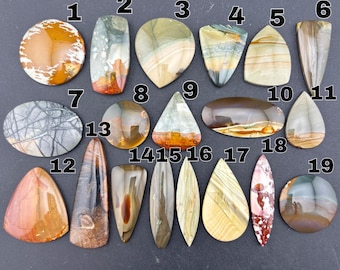 Puede incluir: Diecinueve piedras preciosas cabujón pulidas y multicolores dispuestas en filas, numeradas del 1 al 19. Las piedras presentan varias formas, incluyendo círculos, óvalos, triángulos y gotas, mostrando patrones naturales en tonos marrón, beige, gris y rojo.