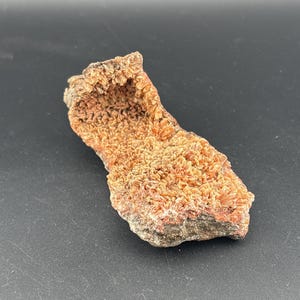 May include: A rough, irregularly shaped rock formation with a textured surface. The rock displays a mix of orange, brown, and gray hues, with a crystalline appearance. The object is set against a dark background.