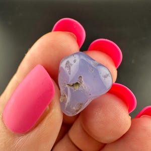 May include: A smooth, rounded, light purple gemstone with small, darker inclusions. The stone is held between fingers with bright pink nail polish.