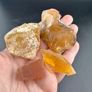 May include: A hand holds several pieces of amber-colored, raw copal resin. The copal pieces vary in size and shape, with some having a rough, textured surface and others appearing more translucent. The resin's color ranges from light yellow to a deeper orange.