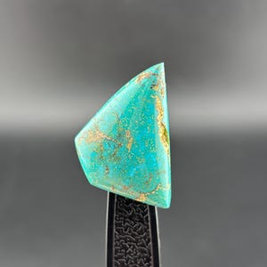 May include: A turquoise gemstone with gold veining, held by a black tool. The stone's polished surface reflects a vibrant blue-green hue. The triangular shape and unique pattern suggest it's for jewellery or collecting. The gemstone is likely measured in centimetres.