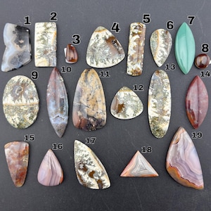 Puede incluir: Una colección de diecinueve piedras preciosas pulidas de diversas formas y tamaños, numeradas del uno al diecinueve. Las piedras presentan diversos patrones, incluyendo inclusiones dendríticas, capas de ágata bandeada y colores sólidos en tonos marrón, gris, blanco y verde.