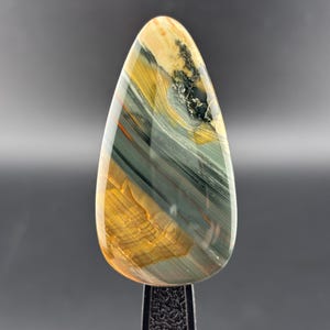 May include: A polished, teardrop-shaped gemstone with a mix of colors. The stone features bands of gold, gray, and black, with hints of red. The gemstone is mounted on a black stand, showcasing its natural patterns and textures.
