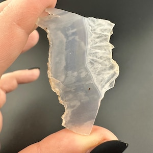 May include: A translucent white agate with a jagged edge and a smooth, polished surface. The agate has a light blue hue and a white band running through it.