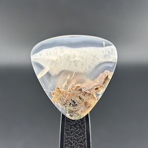 May include: A translucent, triangular-shaped gemstone with a unique pattern. The stone features a white, cloud-like formation at the top, transitioning to a mix of brown and tan earthy textures at the bottom. The gemstone is held by a black clamp.
