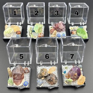 Puede incluir: Siete vitrinas transparentes de acrílico, cada una con una colección única de gemas y minerales coloridos. Las vitrinas están numeradas del 1 al 7. Las gemas incluyen tonos de verde, morado, marrón y amarillo, sobre un lecho de material blanco.