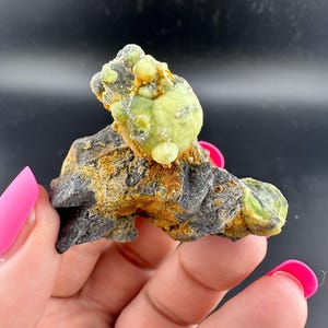 Pode incluir: Um espécime mineral natural com um aglomerado de cristais arredondados verde claro sobre uma matriz rochosa cinza escuro e marrom alaranjado. Os cristais têm uma textura ligeiramente irregular. A aparência geral é áspera e orgânica.