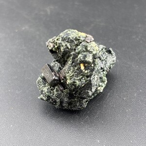 May include: A dark green mineral specimen with several black, geometric crystal formations. The rough surface has a textured appearance, with small, dark crystals and a few lighter-colored spots.