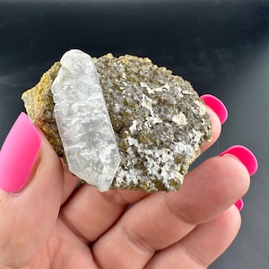 May include: A clear quartz crystal is embedded in a rough, textured rock formation. The rock has a mix of olive green and gray tones, with a crystalline structure. The quartz crystal is translucent and rectangular in shape.