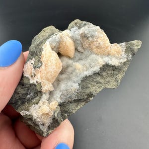 May include: A natural mineral specimen with a dark grey matrix and light tan and white crystal formations. The crystals have a rounded, clustered appearance. The mineral is held by a hand with blue nail polish.