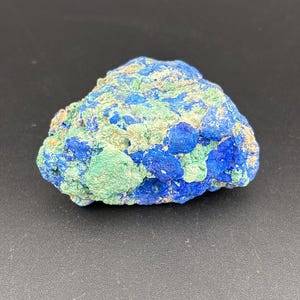 Pode incluir: Um espécime natural de azurite e malaquite. A rocha apresenta uma mistura de azurite azul profundo e malaquite verde, com uma superfície áspera e texturizada. A formação mineral está sobre um fundo escuro.